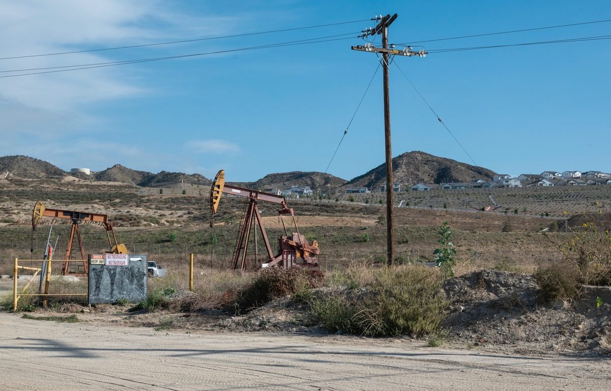 Two pumpjacks on a dry hillside beside a dirt road and a power pole, with brown hills behind.