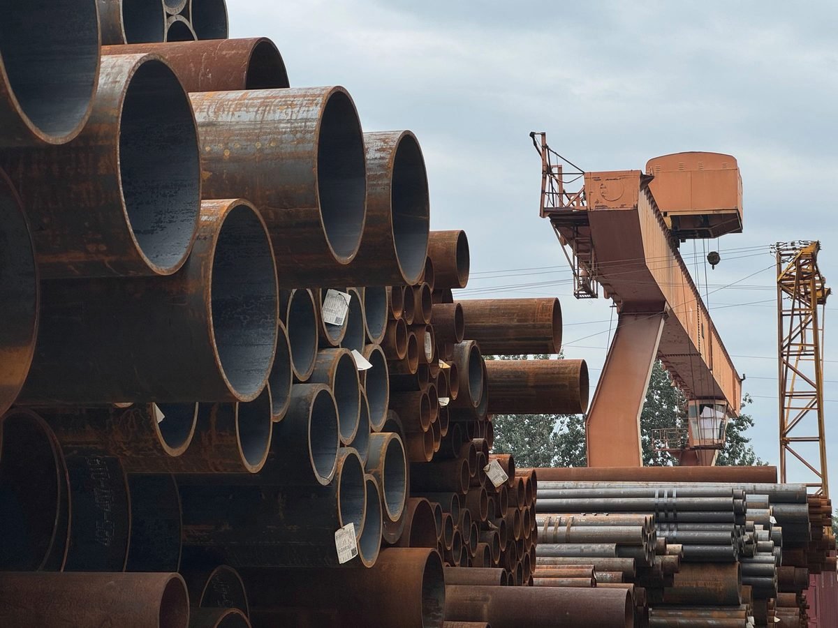 Rusted steel pipe stacked high in a yard, with an orange gantry crane against a grey sky.